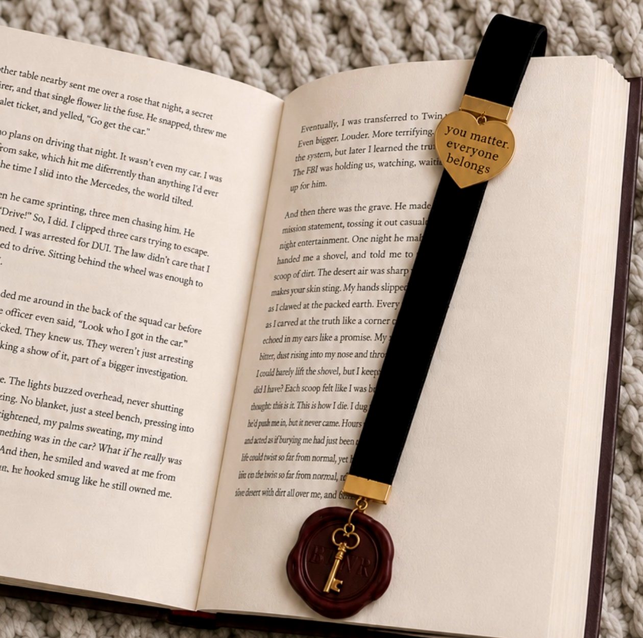 The Signature Velvet Bookmark — black velvet ribbon, a gold heart engraved “you matter. everyone belongs”, and a vintage key pressed in burgundy wax, resting on an open copy of the book